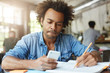 © wayhome.studio  - Concentrated dark-skinned college student with Afro hair doing homework, sitting at canteen table with textbook and copybook, making small break to read text message on his electronic device