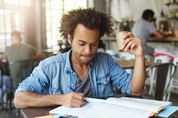 Wall Mural - Indoor shot of serious handsome black male student drinking coffee while working on home assignment, writing down in copybook using pen, looking at notes with focused expression and pursuing lips