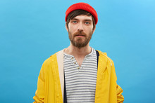 Bearded Sailor Dressed In Red Hat And Yellow Anorak Posing Against Blue Background. Serious Man With Beard Having Blue Charming Eyes Dressed Casually Posing At Studio. Fisherman Going To Angling