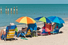 Senior Men And Woman Sitting Under Coloful, Beach Umbrellas Watching Brown Pelicans Standing On Wood Pilings Close To Shore On A Tropical Beach On The Gulf Of Mexico