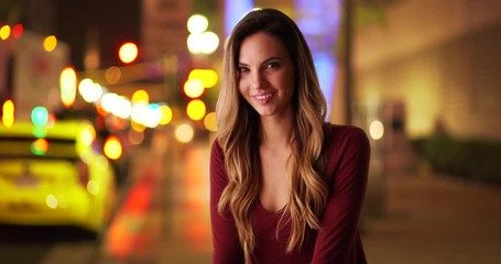Wall Mural - Portrait of attractive female millennial sitting outside downtown at night, smiling at camera. Casual portrait of young Caucasian woman on city street laughing at camera. 4k 