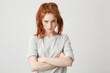 Portrait of resentful young pretty redhead girl looking at camera brutally with crossed arms over white background.