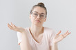 Indoor photo of confused young female wearing trendy hipster eyeglasses isolated on grey background expressing helplessness and indifference with arm gesture, mouth curved in uncertain expression.
