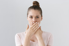Horizontal Photo Of Young European Female Isolated On Gray Background With Expression Of Secrecy And Mistrust As She Is Covering Mouth With Two Hands Not Willing To Disclose Something Important.