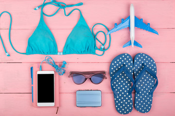 Travel concept - summer women's fashion with blue swimsuit, sunglasses, smart phone, headphones, flip flops, note pad and little airplane and suitcase