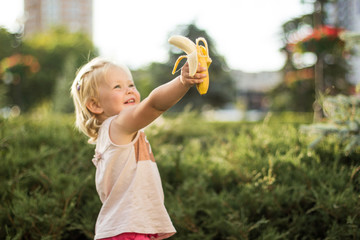 Sticker - The little girl is holding a banana in her hand and laughing. Selective focus. A park.