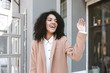 © Anton - Young African American girl smiling and showing hi gesture on street. Cheerful girl standing in beige coat and white shirt. Portrait of pretty lady waving and saying hello