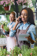 smiling young african american florist holding notebook and talking on smartphone while colleague working behind in flower shop