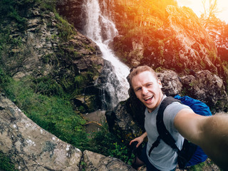 tourist man on a waterfall background holds an action camera and takes a picture of Selfie.