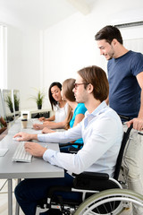 disabled handicapped young male worker on wheelchair with coworker in business office working on a desktop computer