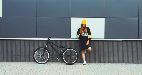 Wall Mural - Young stylish Caucasian female standing near a bicycle against grey wall background, texting on her phone. 4K UHD RAW edited footage