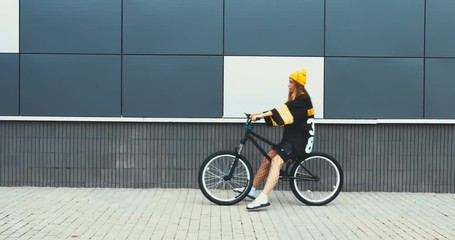 Wall Mural - Young stylish Caucasian female sitting on a bicycle against grey wall background, texting on her phone. 4K UHD RAW edited footage