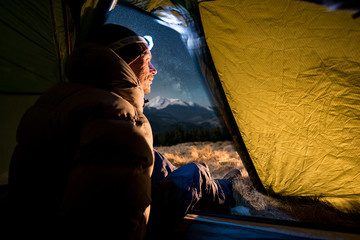 Wall Mural - View from inside a tent on the male hiker have a rest in his camping at night. Man with a headlamp sitting in the tent near campfire