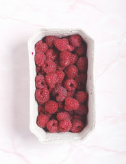 Fresh raspberries in a container on pink marble background. Seasonal summer fruit. Flat lay. Top view.