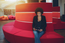 Young Serious Afro American Girl With Curly Hair Is Sitting On Red Curved Sofa In Chillout Office Space And Reading E-mail From Her Boss About Future Meeting With Copy Space Place For Text Or Logo