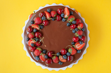 Wall Mural - Birthday cake in chocolate with strawberries, blueberries and cherry on yellow background. Top view. Picture for a menu or a confectionery catalog.