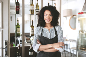 Beautiful African American girl in apron standing with arms folded in restaurant. Young girl with dark curly hair standing in apron at cafe. Portrait of smiling waitress wearing uniform