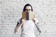 Pretty hipster tattooed woman poses in white t-shirt with book in hands, isolated on white brick wall, Hold a book in hand, space for design layout