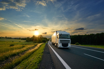 Wall Mural - white truck driving on the asphalt road in rural landscape at sunset