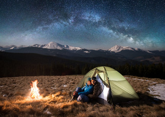 Wall Mural - Young couple tourists having a rest in the camping at night, sitting near campfire, looking to the camera under beautiful starry sky and milky way. On the background snow-covered mountains