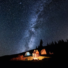 Wall Mural - Night camping in the mountains. Couple tourists have a rest at a campfire near two tents under beautiful night sky full of stars and milky way. Long exposure