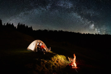 Wall Mural - Night camping in the mountains. Couple tourists have a rest in the illuminated tent near campfire under beautiful night sky full of stars and milky way. Low light