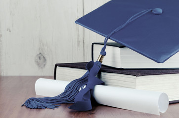 a blue graduation cap resting on two books with a diploma tied with blue ribbon on a wooden backgrou