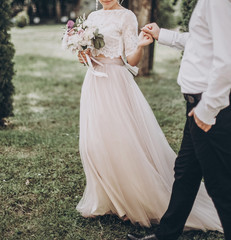 Wall Mural - stylish wedding bride and groom walking in sunny park holding hands. modern couple holding fashionable bouquet, woman smiling. fine art wedding photo, romantic tender moment