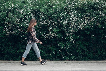Hipster Woman In Sunglasses Walking On Background Of Blooming Bush With White Flowers Of Spirea. Boho Girl Outing In Floral And Denim Modern Clothes In Summer City Street. Space For Text