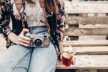 Wall Mural - stylish hipster woman holding lemonade and old photo camera. boho girl in denim and bohemian clothes, holding cocktail sitting on wooden bench at street food festival. summertime