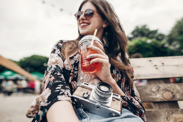 happy stylish hipster woman holding lemonade. cool boho girl in denim and bohemian clothes, with cocktail and old camera at street food festival. summer vacation travel