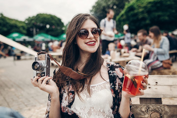 Wall Mural - stylish hipster woman in sunglasses with red lips holding lemonade and old photo camera. boho girl holding cocktail and smiling at street food festival. summertime. summer  vacation