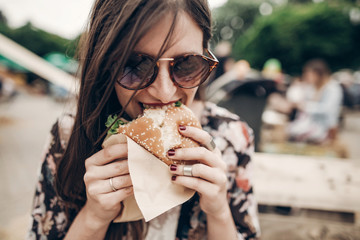 Wall Mural - stylish hipster woman eating juicy burger. boho girl biting yummy cheeseburger, smiling at street food festival. summertime. summer vacation travel picnic. space for text