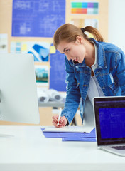 Two young woman standing near desk with instruments, plan and laptop.