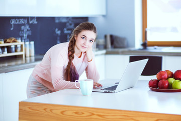 Young woman sits at the kitchen table using a laptop and talking on a cell phone.