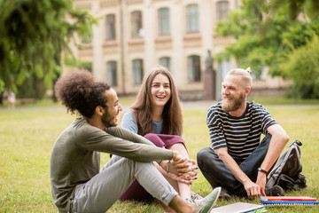 Happy laughing students at University garden. Three studens having lively conversation sitting on grass.