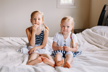 Wall Mural - Portrait of two cute adorable little red-haired blonde Caucasian girls sisters sitting together on bed at home. Siblings looking in camera. Happy lifestyle childhood concept.