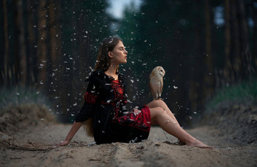 girl sits on forest road with owl on her knees among flying fluff .