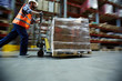 Blurred motion shot of warehouse worker wearing hardhat and reflective jacket pushing moving cart with boxes along isle between tall racks