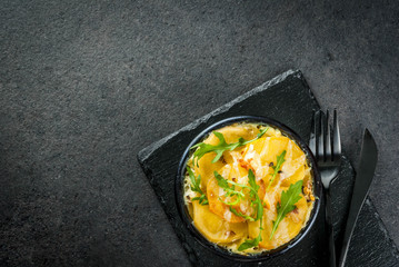 Casserole. French cuisine. Homemade potato gratin in a bowl for baking. On a black stone background. With leaves of fresh arugula for decoration. Top view copy space