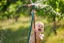 A Boy Play With Water Sprinkler In The Summer Garden