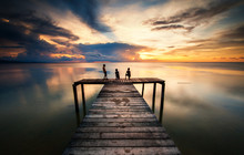 Wooden Jetty During Sunset With Reflection. Three Unknown Sihleoutte Of  Kids Appear On The Image.  Soft Focus Due To Long Expose.