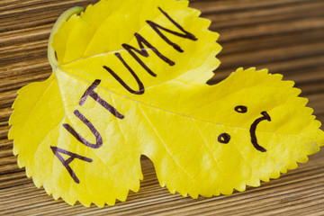 Autumn yellow leaf with inscription Autumn and a picture of unhappy face on the wooden background. Selective focus, closeup