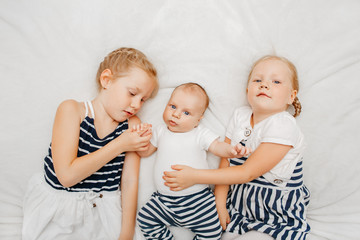 Wall Mural - Lifestyle portrait of cute white Caucasian girls sisters holding little baby, lying on bed blanket indoors. Older siblings with younger brother sister newborn. Family love bonding together concept.