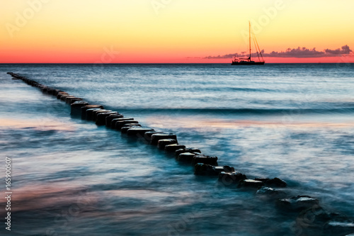 Wooden groynes and sailing boat. Red sunset and long exposure. Poster