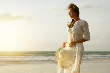 Wall Mural - Woman wearing beautiful white dress is walking on the beach during sunset