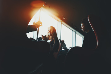 Wall Mural - Silhouettes of two business women in dark office settings sitting on armchairs with their gadgets and having lively discussion with their third colleague during work meeting near window