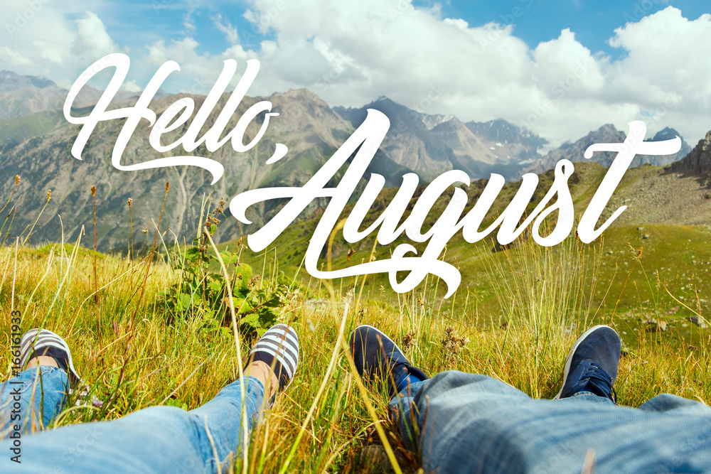 Hello august text with couple's legs together on view of the green valley and high mountains. Nature calendar background. Travel, vacation, holiday, unity with nature, adventure concept. - obrazy, fototapety, plakaty 