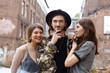 © shurkin_son - Attractive young European man wearing trendy hat and camouflage jacket relaxing in urban setting with his two girlfriends, enjoying good company. Three cheerful teenage friends having fun outdoor