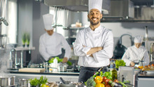 Famous Chef Of A Big Restaurant Crosses Arms And Smiles In A Modern Kitchen. His Staff In Working In The Background.